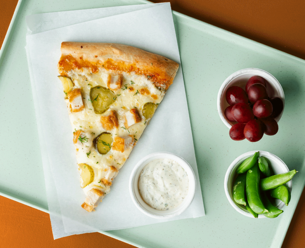 Slice of pickled pizza with ranch dipping sauce on a blue tray with grapes and snap peas